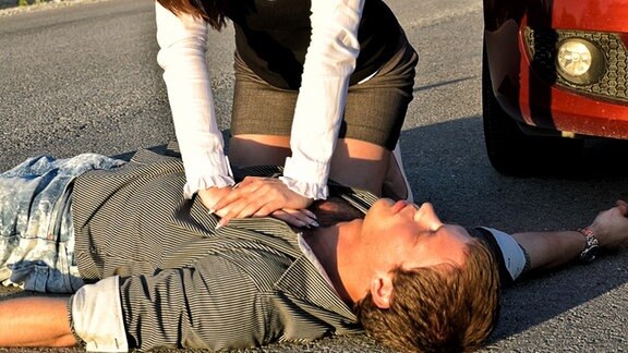 Eine Frau kniet neben einem jungen Mann, der leblos auf der Straße liegt, und führt eine Herzdruckmassage durch.